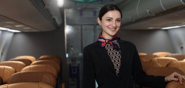 Uçuş görevlisi ya da hostes. Dost canlısı bir uçuş görevlisi, bir uçak kabininin içinde güvenle durur. Profesyonel konukseverlik ve zarif üniforma. Havacılık hizmeti seyahat havayolu hostesi.