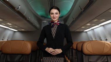 Uçuş görevlisi ya da hostes. Dost canlısı bir uçuş görevlisi, bir uçak kabininin içinde güvenle durur. Profesyonel konukseverlik ve zarif üniforma. Havacılık hizmeti seyahat havayolu hostesi.