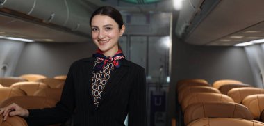 Hostes. Üniformalı gülen uçuş görevlisi modern bir uçağa binen yolcuları karşılıyor ve onlara rehberlik ediyor. Arkadaş canlısı havayolu personeli profesyonel hizmet ve konforlu uçuş deneyimi sunuyor