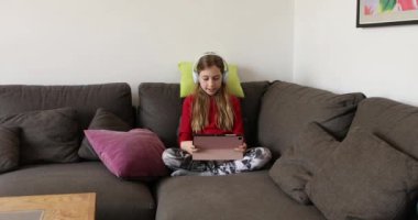 Kulaklıklı küçük bir kız evdeki kanepede oturmuş tabletiyle oynuyor.