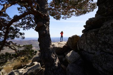 İspanya, Madrid 'deki Guadarrama Ulusal Parkı' ndaki Cercedilla Dağı 'ndan ufuktaki vadide dürbünle ağaçların arasındaki bir kayanın tepesindeki kadın.