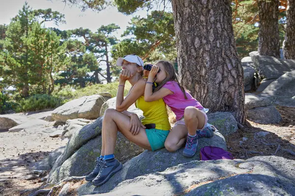 Anne ve kız yürüyüşçüler ormandaki bir kayanın üzerinde dinleniyorlar. Kız dürbünle bakıyor. İspanya, Madrid 'deki Guadarrama Ulusal Parkı dağlarında.