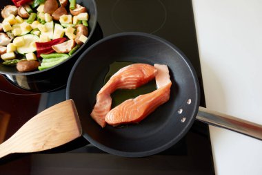 Endüksiyon fırınında iki tava pişiriliyor: biri iki parça somon balığı filetosu ve diğeri de yeşil ve kırmızı biber, kabak, mantar, yabani kuşkonmaz ve elma gibi kesilmiş sebzelerle.