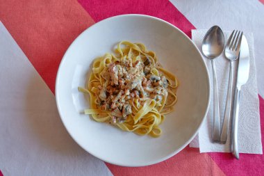 Pastırmalı, mantarlı, soğanlı, kremalı ve fesleğenli tagliatelle carbonara makarnası. Çatal, bıçak, kaşık ve kağıt peçetenin yanında renkli bir masa örtüsünün üzerinde.