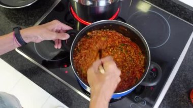 Bolonez sosu pişiren, kızarmış domates, kıyma, kabak, mantar dilimleri, ahşap kaşıkla doğranmış soğan karıştıran, evdeki elektrikli ocakta paslanmaz tencerenin yanında bir kadının elleri. 