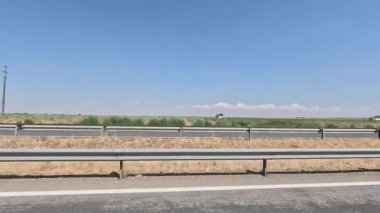 Parlak, bulutsuz bir gökyüzü altında yeşil tarım arazileri ve kuru bitki örtüsüyle çevrili metal bariyerli sakin bir İspanyol otoyolunun kenar görüntüsü, sakin kırsal manzarayı gözler önüne seriyor.
