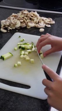 Evdeki mutfak tezgahında, dilimlenmiş mantarların yanında beyaz bir kesme tahtasıyla kabak kesen bir kızın dikey görüntüsü.