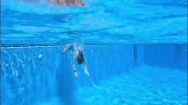 Bir kız havuza balıklama atlıyor ve suyun altında parlak, berrak bir mavi arka planla yüzüyor, yaklaşıyor ve gülümsüyor.