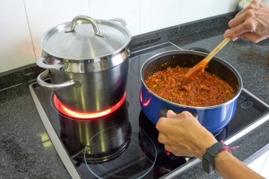 Evde elektrikli fırında makarna hazırlayan iki tencere. Bir tencere domates soslu, diğeri de makarnalı ve parlak kırmızı ocaklı.