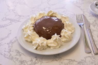 Beyaz bir tabakta çırpılmış krema kıvrımlarıyla çevrili zengin çikolatayla kaplı yozlaşmış bir tatlı. Yanında bir çatal ve bıçak duruyor, bu zevkli ziyafetin tadını çıkarmaya hazır.