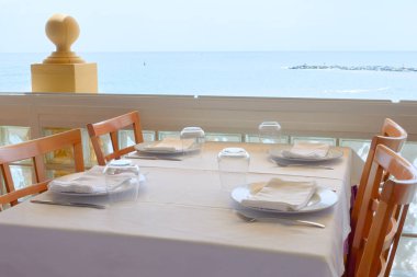 An elegant restaurant table set with white linens and place settings, overlooking the ocean through large windows. Wooden chairs complement the coastal dining atmosphere clipart