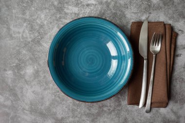 Gümüş çatal ve bıçaklı boş turkuaz seramik tabak, gri beton veya taş zemin üzerinde kahverengi peçete, fotokopi alanı manzaralı.
