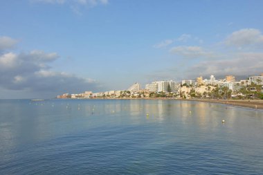 Benalmadena Costa 'nın sakin sahil manzarası, Malaga köyü, Endülüs, İspanya. Mavi su ve sahil kenarındaki binalar parçalı bulutlu bir gökyüzünün altında, sakin bir Akdeniz kıyısı gösteriyor.