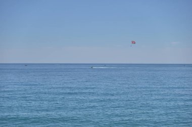 Berrak bir gökyüzü altında sakin mavi sulara sahip sakin bir deniz manzarası. Uzak bir deniz paraşütü aktivitesi küçük kırmızı bir nokta gibi görünür. Huzurlu bir Akdeniz yaz atmosferi yaratır.