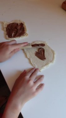 Küçük bir kızın ev yapımı tarçın ruloları yapmak için bıçakla ekmek dilimlerine çikolata ve fındık serpiştirdiği dikey video.