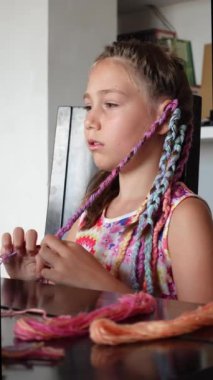 Saçlarını renkli yünle ören genç bir kız oturma odasında çok yetenekli bir kuaför gibi oturuyor. Dikey video