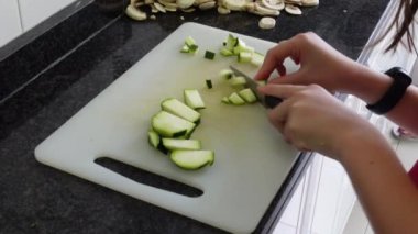Evdeki mutfak tezgahında bir yığın doğranmış mantarın yanında beyaz kesme tahtasına bıçakla kabak kesen bir kızın elleri.
