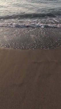 Okyanus dalgalarının kumlu bir sahilde yavaşça yuvarlanışının yakın görüntüsü. Köpüklü beyaz su, dalgalar dalgalanırken ve kıyı şeridi boyunca akarken kahverengi ıslak kumla çelişiyor. Yavaş çekim dikey video
