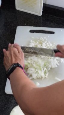Evdeki mutfakta beyaz bir kesme tahtasıyla soğan doğrayan bir kadının dikey görüntüsü.