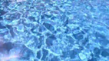 Genç bir kız açık mavi bir havuzda yüzüyor, koyu kırmızı bir mayo giyiyor. Yüzeyin dışından bakıldığında güneş ışığı vücudunda ve havuz zemininde dalgalanma izleri yaratıyor.