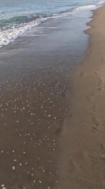 Kumsalda yürüyen dikey bir video. Deniz dalgaları kahverengi kumlara çarpıyor. Bazı ayak izleri var.