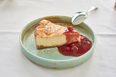 Restoran masasının beyaz masa örtüsünde yeşil tabakta ahududu reçelli bir porsiyon cheesecake. Tabakta çelik bir kaşık duruyor.