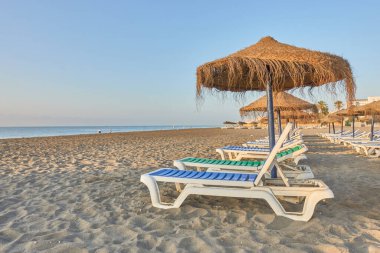Torremolinos, Malaga 'daki Los Alamos plajının ön sırasındaki kumsalda güneşlik ve saman şemsiyesi. Gün doğumunda ufuk çizgisi ve açık gökyüzü ile Akdeniz manzarası. Endülüs, İspanya