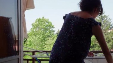 Ciddi şekilde odaklanmış çekici 40 'lı yaşlarda Latin kökenli, gözlüklü bir kadın teras balkonunda temiz çamaşır asıyor.