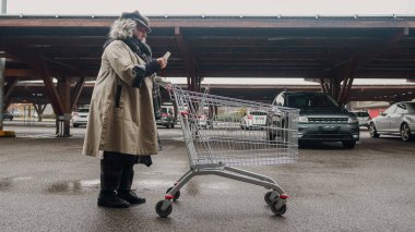 İspanyol genç kadın el arabası itiyor. El yazısı okuyor. Büyük bir süpermarketin otoparkında alışveriş listesi hazırlıyor.