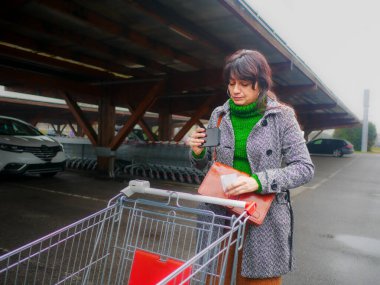 Latin Amerikalı genç bir kadın market otoparkında alışveriş listesi okuyarak el arabası itiyor.