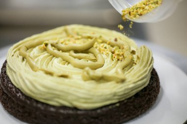 Pasta şefi tasarımcısı, mutfak laboratuarındaki katmanlı çikolatalı kekin üzerine fıstık kreması ve çikolata serpiştiriyor.