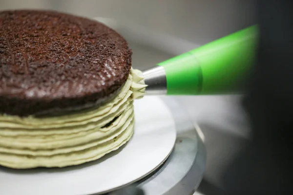 cake chef designer using pistachio cream filling piping bag on layered dark chocolate cake at kitchen lab