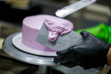 Pasta şefi pürüzsüz leylak pembesi kremalı pasta yapmak için spatula kazıyıcısı kullanıyor. Doğum günü pastası hazırlığı.