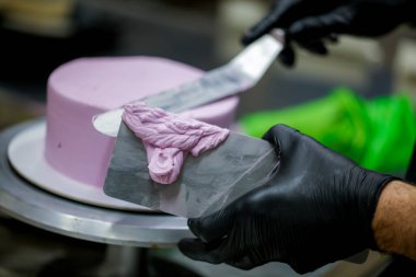 Pasta şefi pürüzsüz leylak pembesi kremalı pasta yapmak için spatula kazıyıcısı kullanıyor. Doğum günü pastası hazırlığı.