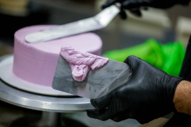 Pasta şefi pürüzsüz leylak pembesi kremalı pasta yapmak için spatula kazıyıcısı kullanıyor. Doğum günü pastası hazırlığı.