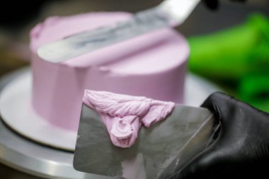 Pasta şefi pürüzsüz leylak pembesi kremalı pasta yapmak için spatula kazıyıcısı kullanıyor. Doğum günü pastası hazırlığı.