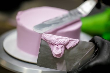 Pasta şefi pürüzsüz leylak pembesi kremalı pasta yapmak için spatula kazıyıcısı kullanıyor. Doğum günü pastası hazırlığı.