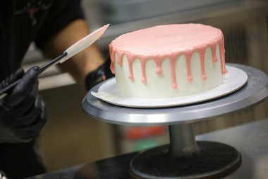 Pasta şefi spatulayı kullanarak beyaz kremalı kekin üzerine pembe damlatarak mutfak laboratuarında doğum günü kutlaması yapıyor.