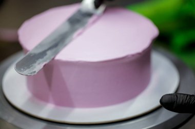 Pasta şefi pürüzsüz leylak pembesi kremalı pasta yapmak için spatula kazıyıcısı kullanıyor. Doğum günü pastası hazırlığı.
