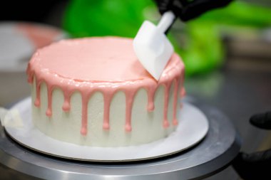 Pasta şefi spatulayı kullanarak beyaz kremalı kekin üzerine pembe damlatarak mutfak laboratuarında doğum günü kutlaması yapıyor.