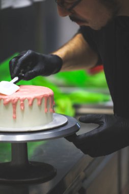 Pasta şefi spatulayı kullanarak beyaz kremalı kekin üzerine pembe damlatarak mutfak laboratuarında doğum günü kutlaması yapıyor.