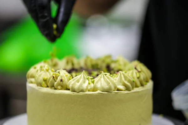 pastry designer sprinkling the top of green layered frosted chocolate cake with small pistacchio and dark chocolate pieces