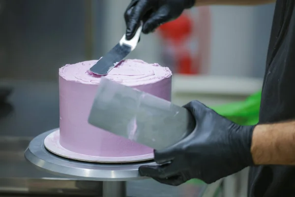 pastry chef using spatula and scraper smoothing levelling layered lilac buttercream frosted cake - wedding cake preparation
