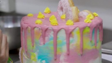 Pasta tasarımcısı en üstteki 4K görüntüde pinl sarı deniz mavisi boru kremi ve donut ile süslenmiş kişniş kaplı bir pasta hazırlıyor.