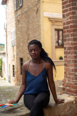 Rahat, genç, siyah bir kadın oturuyor. Şık bir model. Örgü örme saç stili. Seçici dış mekan odaklı özgüvenli Afrikalı kadın moda tarzı, mutluluk konsepti. Açık havada..