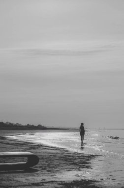 Genç kadın kumsalda şık kıyafetler içinde poz veriyor. Arka planda güzel deniz manzarası. Esmer, uzun boylu, esmer ve zayıf. Yaz, gün batımı. Siyah beyaz resim.