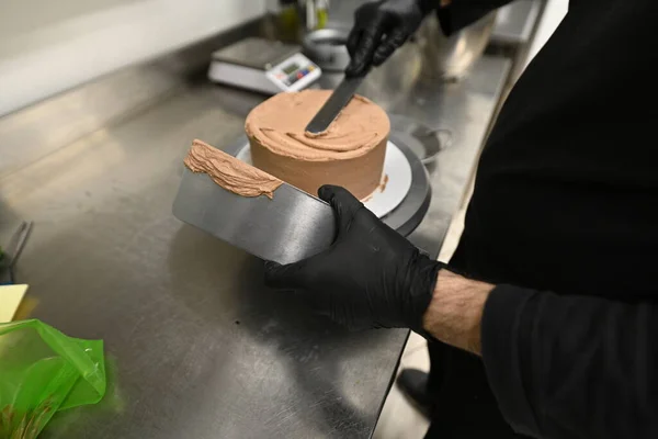 pastry chef smoothing cream on a frosted layered dark chocolate cake in professional kitchen high res image