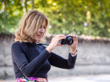 Kafkasyalı 70 'li yaşlarda formda, mutlu bayan. Parkta fotoğraf makinesiyle rahatla. Çiçekli etek ve siyah uzun kollu gömlek giy.