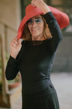 Kırmızı gözlüklü, siyah elbiseli ve dans ayakkabılı olgun bir kadın bir fotoğrafa poz verirken kendine güvenen seksi bir poz veriyor..