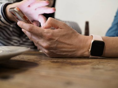 Akıllı saat takan bir kadın yemek yerken telefonunu kontrol ediyor, elleri ahşap bir masada dinleniyor, teknolojiyi ve sosyal etkileşimi iç mekanda harmanlıyor.